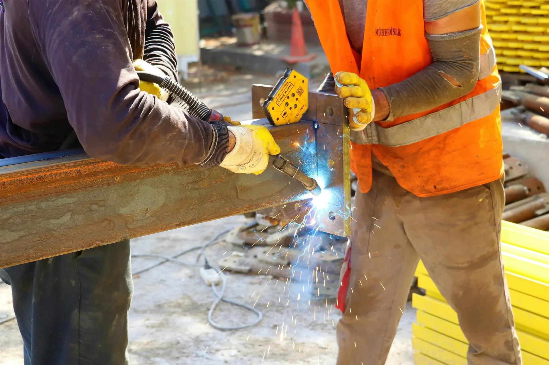Two workers in safety gear welding metal at a construction site, emitting sparks.